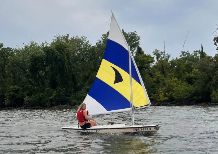 A person wearing a red life jacket sails a small boat with a blue, yellow, and white sail on calm water, with trees and greenery in the background under a cloudy sky.