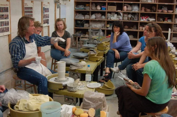 A group of people sit around a pottery wheel watching a person in an apron demonstrate pottery techniques in a studio filled with shelves of clay and ceramics supplies.
