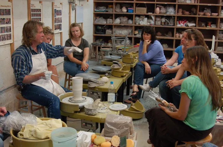 A group of people sit around a pottery wheel watching a person in an apron demonstrate pottery techniques in a studio filled with shelves of clay and ceramics supplies.