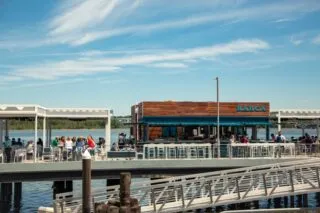 People dine at an outdoor waterfront restaurant with a wooden building labeled BARCA under a blue sky. The restaurant is situated on a pier over the water, with tables and seating areas on both sides.