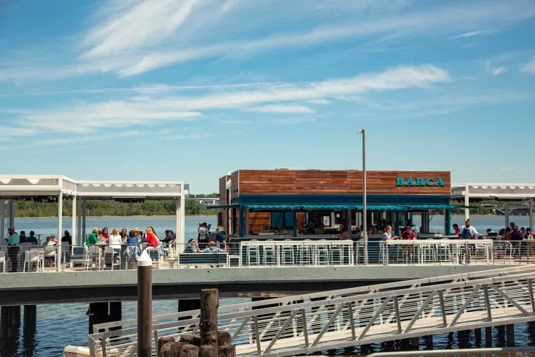 People dine at an outdoor waterfront restaurant with a wooden building labeled BARCA under a blue sky. The restaurant is situated on a pier over the water, with tables and seating areas on both sides.