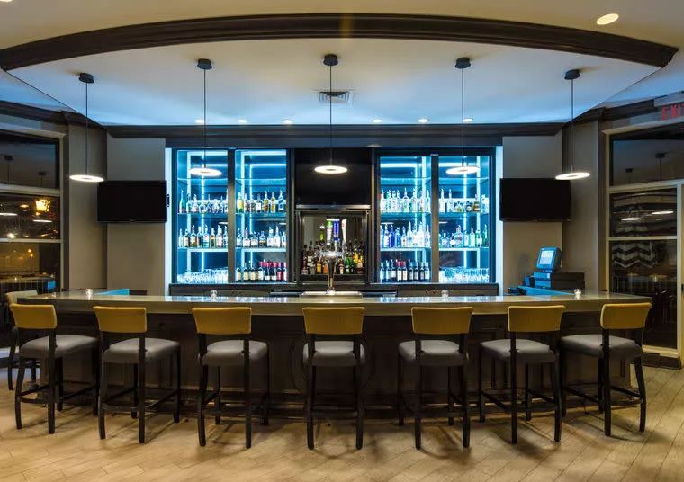 A modern bar with a long counter, high chairs, and illuminated shelves displaying various bottles of alcohol. There are two wall-mounted TVs, pendant lights above, and a cash register on the right side of the counter.
