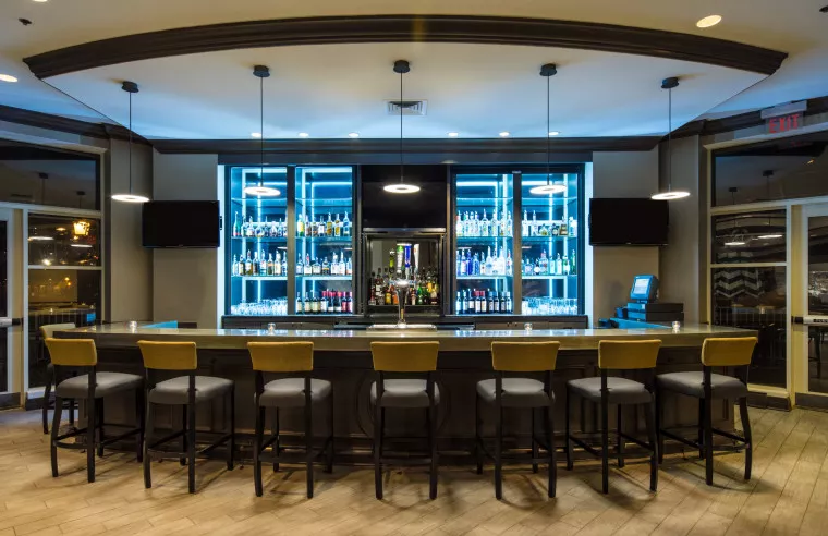 A modern, well-lit bar with high chairs lined along a counter, illuminated shelves displaying various bottles of alcohol, and two flat-screen TVs mounted on either side. The space has a clean, contemporary design.