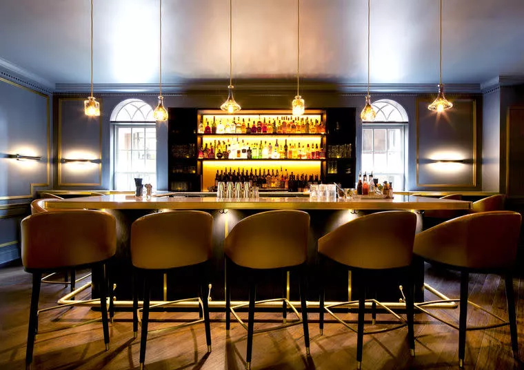 A modern, elegant bar with six tan barstools, pendant lights hanging above, and a backlit shelf displaying various bottles of liquor. The bar has a sleek, inviting atmosphere with warm lighting and large arched windows.