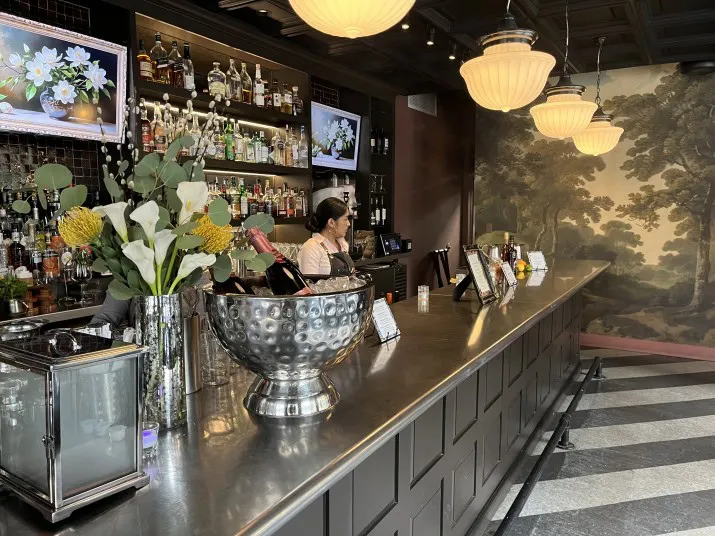 A bartender stands behind a modern bar with a metal countertop, floral arrangement, bottles of alcohol, and large hanging lights. A mural of trees decorates the wall in the background.