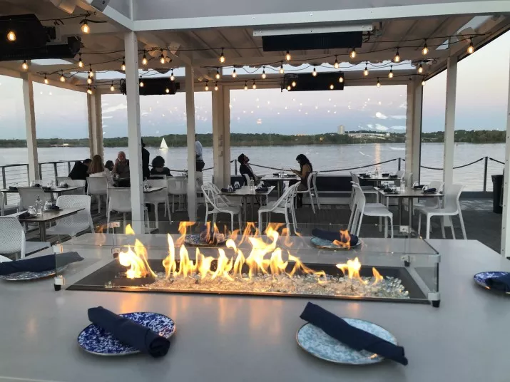 A waterfront patio restaurant at sunset with a rectangular fire pit in the center, blue and white plates with navy napkins on tables, string lights overhead, and diners seated by the railing overlooking the water.