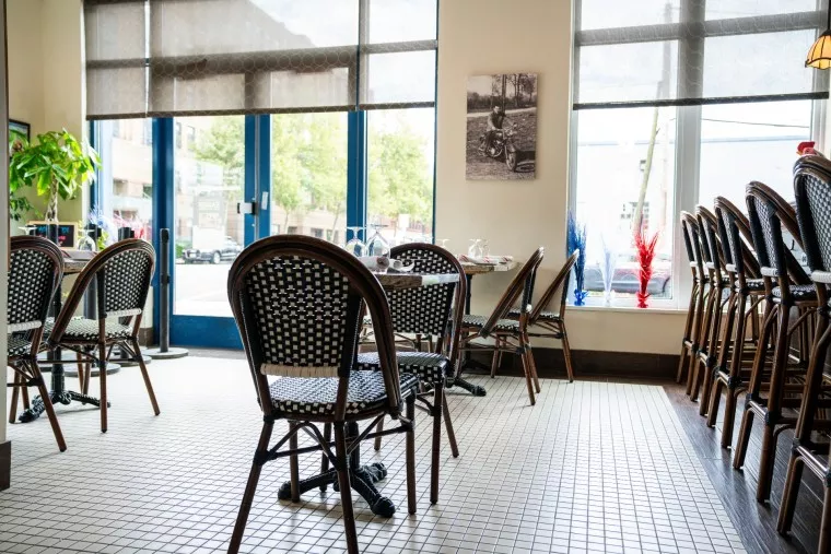 A bright, empty cafe with checkered bistro chairs, tables set for dining, large windows letting in natural light, and a black-and-white photo on the wall. Some chairs are stacked near the window.