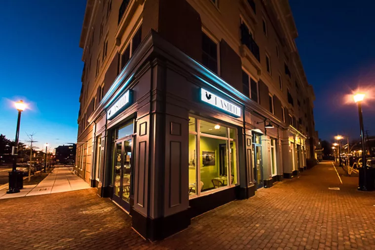 A well-lit corner storefront with large windows and a sign reading VAN SMITH on a brick walkway at dusk, with street lamps and a multi-story building above.