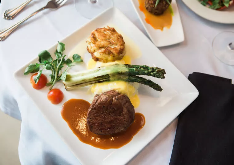 A plated meal featuring a steak with brown sauce, two spears of asparagus, a twice-baked potato, halved cherry tomatoes, greens, and a creamy yellow sauce, served on a white square plate with silverware nearby.