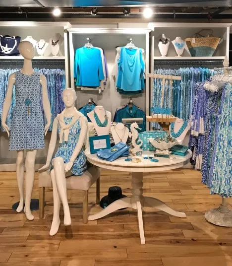 A boutique display features two mannequins in blue-patterned dresses, a round table with jewelry and accessories, and racks of blue and turquoise clothing against a wooden floor and white shelving.