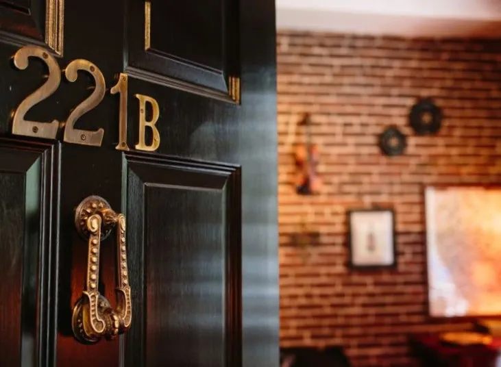 A black door with brass numbers “221B” and a decorative brass knocker is partially open, revealing a cozy interior with exposed brick walls and framed artwork.