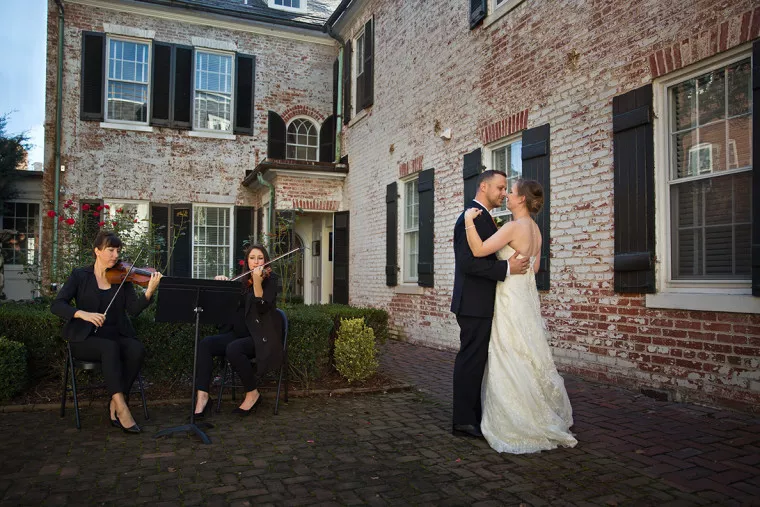 A bride and groom dance together in an outdoor courtyard, while two musicians seated nearby play string instruments. The scene is set against a brick building with black shutters and greenery.