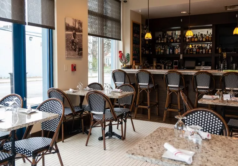 Cozy restaurant interior with empty black-and-white woven chairs, marble tables set with glasses and napkins, tiled floor, and a bar area lined with bottles and high chairs. Large windows let in natural light.
