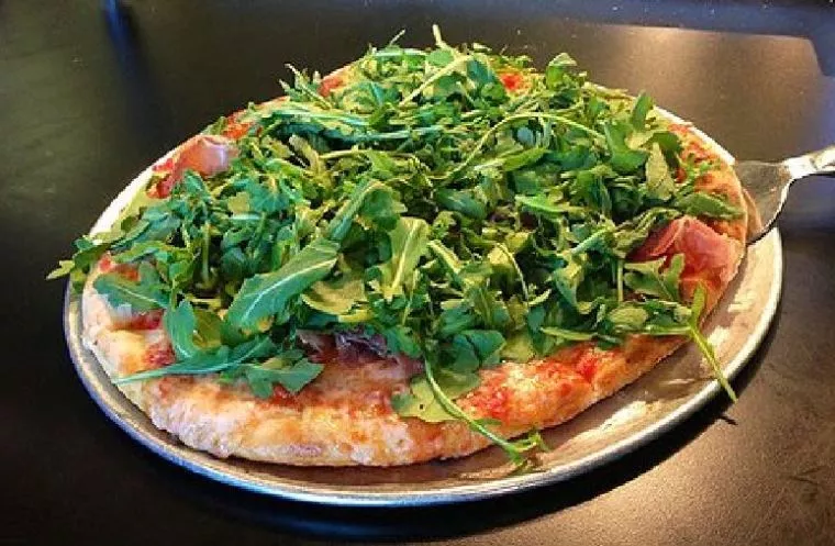 A pizza topped with a generous amount of fresh arugula sits on a metal tray, with some tomato sauce and melted cheese visible beneath the greens.