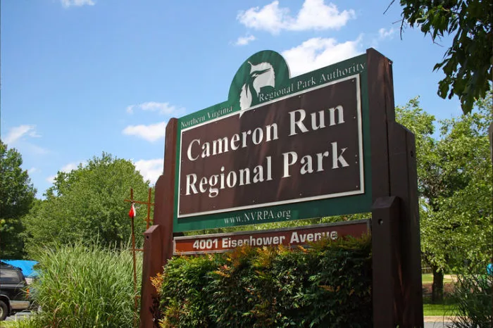 A large brown and green sign reads Cameron Run Regional Park with smaller text for the Northern Virginia Regional Park Authority and the address 4001 Eisenhower Avenue. Trees and greenery surround the sign.