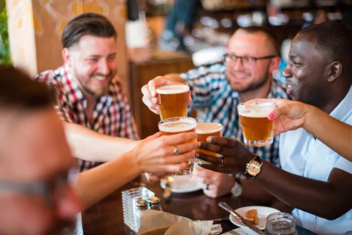 A group of friends sitting at a table in a bar clinking glasses of beer, smiling and enjoying each other’s company.