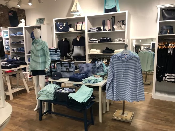 A clothing store displays neatly folded shirts, sweaters, and tops in blue and green tones. A mannequin wears a striped shirt and shorts. Shelves and racks are organized, with accessories and decor adding a nautical theme.