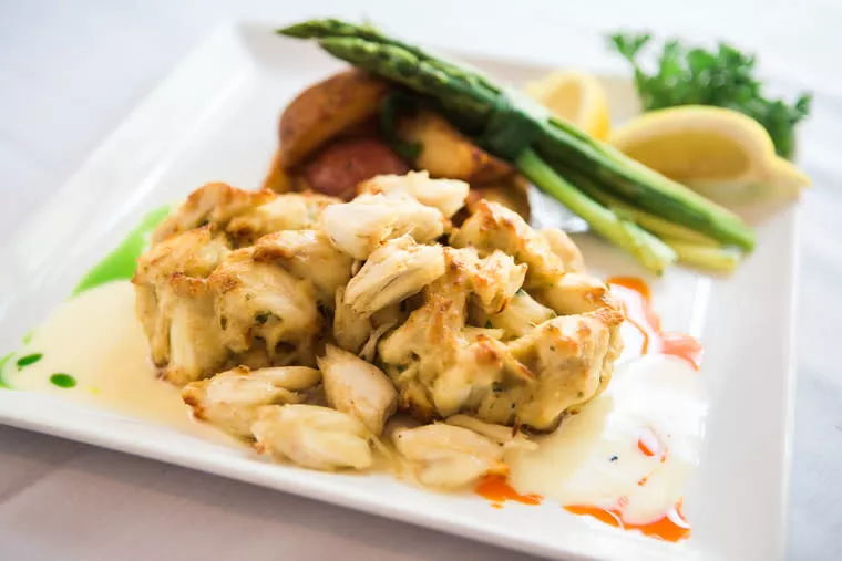 A plate with a crab cake topped with creamy sauce, garnished with green herbs. On the side are roasted potatoes, asparagus spears, lemon wedges, and colorful sauce drizzles on a white rectangular plate.