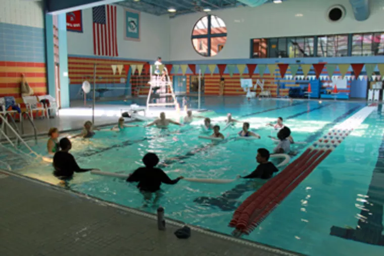 A group of people participate in a water aerobics class in an indoor swimming pool, guided by an instructor on a lifeguard stand. The pool is surrounded by colorful flags and large windows.
