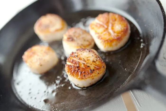 Five seared scallops cooking in a black cast iron skillet, showing a golden-brown crust and a bit of oil sizzling around them.