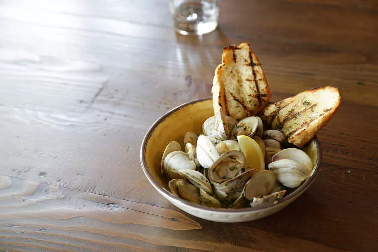 A bowl of steamed clams garnished with herbs and served with lemon wedges and two slices of grilled bread sits on a wooden table. A glass of water is blurred in the background.