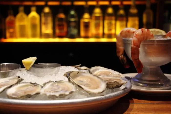 A platter of oysters on ice with lemon and sauce sits next to a metal bowl filled with shrimp and lemon wedges. Bottles of alcohol are blurred in the background, creating a warm ambiance.