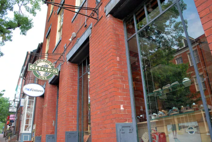A red brick building with large glass windows displays shoes inside. Signs for Comfort One Shoes and ProCentures hang above the sidewalk. Trees and reflections of buildings are visible outside.