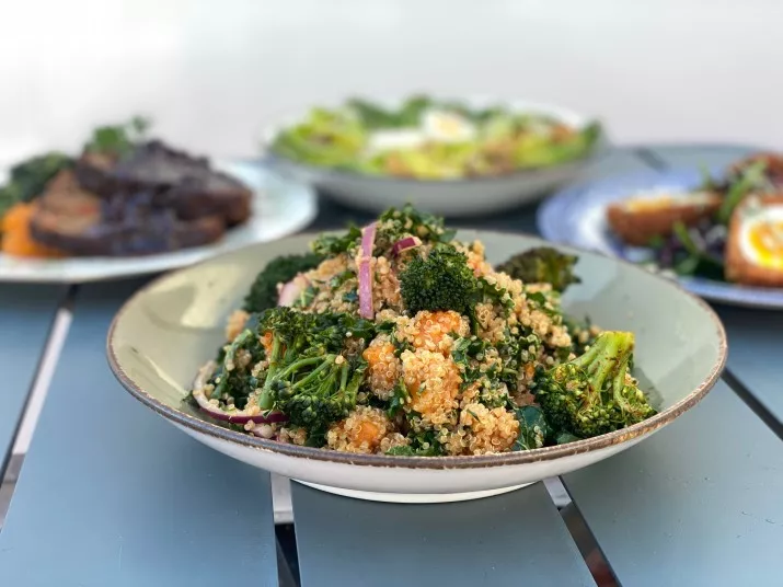 A bowl of quinoa salad with broccoli, red onions, and herbs sits on a table, with plates of various foods blurred in the background.
