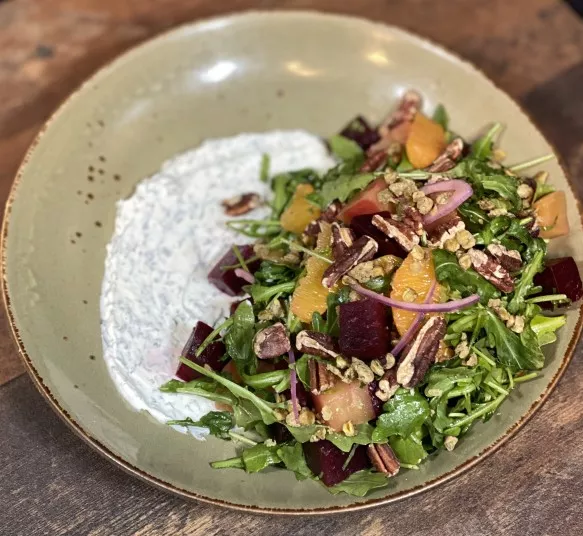 A salad with arugula, beets, orange segments, red onions, pecans, and grains is served on a green plate with a creamy herb dressing spread on one side. The plate rests on a wooden surface.