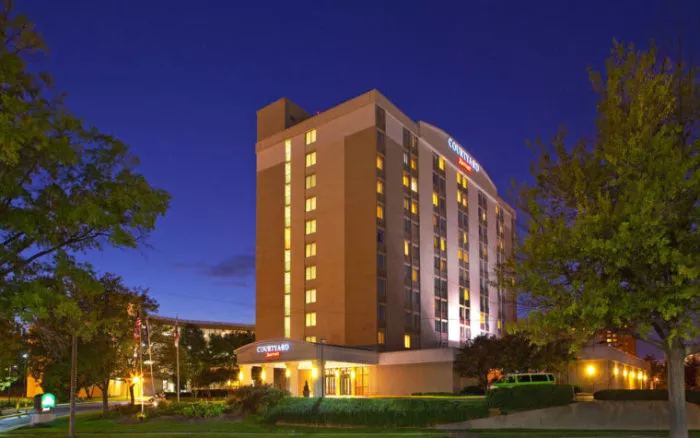 A well-lit, multi-story Courtyard by Marriott hotel at dusk, surrounded by trees and greenery, with lights glowing from the windows and a dark blue evening sky in the background.