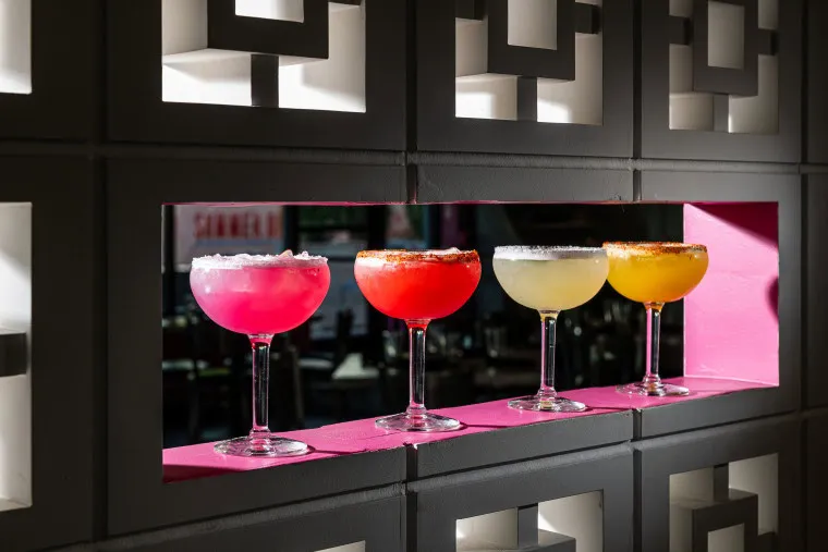 Four colorful cocktails in margarita glasses are lined up on a pink shelf set into a gray decorative wall, each drink displaying a different vibrant hue: pink, red, yellow, and orange.