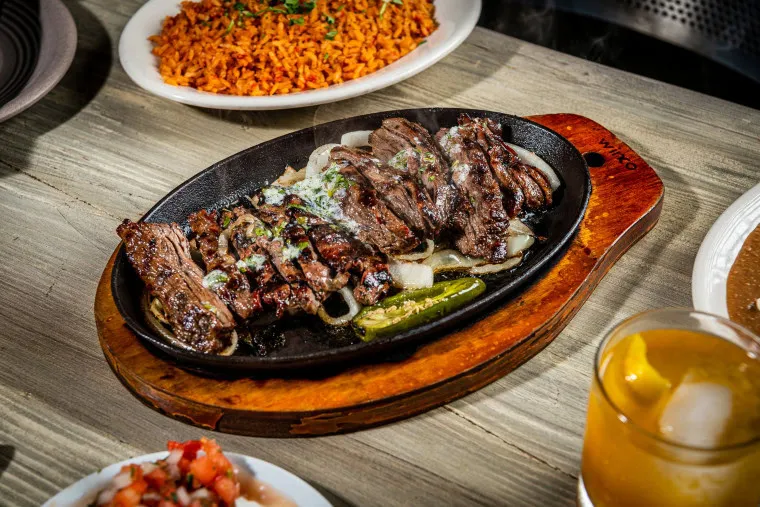 A sizzling platter of grilled steak served over onions and jalapeños, accompanied by a plate of Mexican rice, a glass of drink, and a dish of pico de gallo on a wooden table.