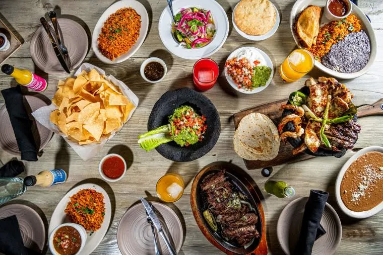 Colorful Mexican feast on a wooden table, featuring grilled meats, guacamole, rice, beans, salsa, chips, pickled onions, tortillas, and assorted drinks arranged around empty plates and utensils.
