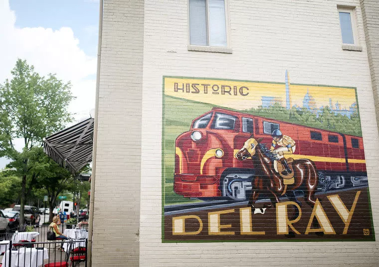 A mural on a brick wall depicts a red train, a horse with a rider, and the words Historic Del Ray, with a city skyline and trees in the background. Some people are seated outside near the building.