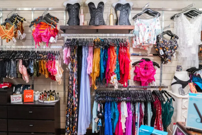 A display of colorful lingerie, corsets, and nightwear hangs on racks and shelves in a store. Various fabrics and patterns are visible, along with accessories and a few small boxes on a nearby dresser.