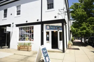 A white brick building with a sign and board reading Now Open, large windows, black trim, and star decorations, stands on a sunny street corner with a tree and sidewalk nearby.
