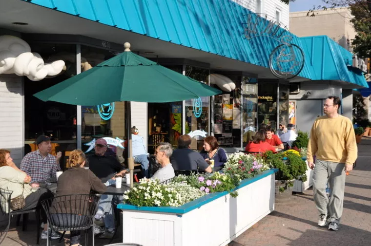 People are dining outdoors at a café with blue awnings, sitting at tables near planters filled with flowers. A man walks past on the sidewalk. The scene looks bright and lively.