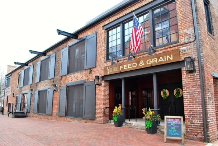 A brick building with dark shutters, an American flag, and a sign reading Virtue Feed & Grain above the entrance. Potted plants and a chalkboard menu stand at the doorway; holiday wreaths hang on the doors.