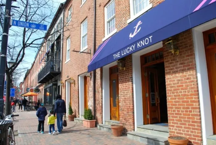 A brick building with a blue awning reading “The Lucky Knot,” located on a sidewalk in a shopping district. People, including children, walk along the street lined with shops and outdoor seating.