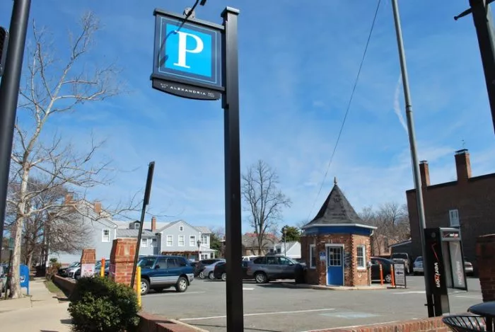 A parking lot with several cars, a blue P parking sign, a small brick booth with a pointed roof, and residential buildings in the background under a clear blue sky.