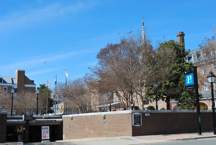 A brick parking garage entrance with a sign showing rates, surrounded by trees and historic brick buildings, with flags and a church steeple visible in the background under a clear blue sky.