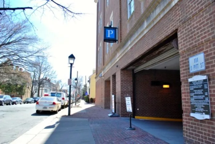 Parking garage entrance with P sign on a brick building at 111 S Pitt St. Cars parked along the street, trees without leaves, and a sidewalk with a lamppost are visible.