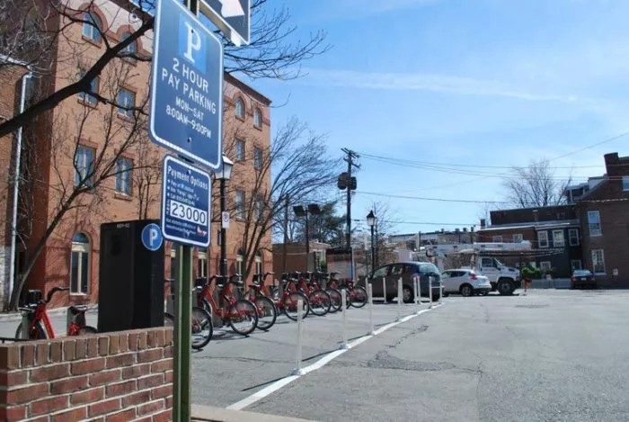 A parking lot with a 2 Hour Pay Parking sign, several parked cars, a row of rental bicycles, and surrounding brick buildings under a clear blue sky.