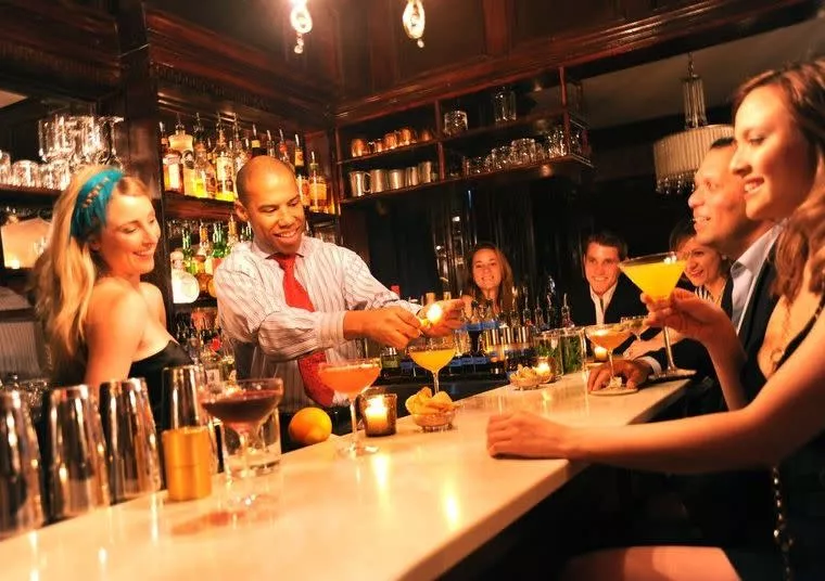 A bartender prepares cocktails for a group of smiling people seated at a bar, holding colorful drinks and enjoying a lively, upscale atmosphere with shelves of liquor bottles in the background.