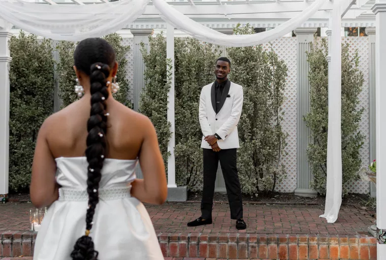 A woman in a white dress with a long braided ponytail faces a man in a white jacket and black pants, who stands smiling under white drapery outdoors with greenery and columns in the background.