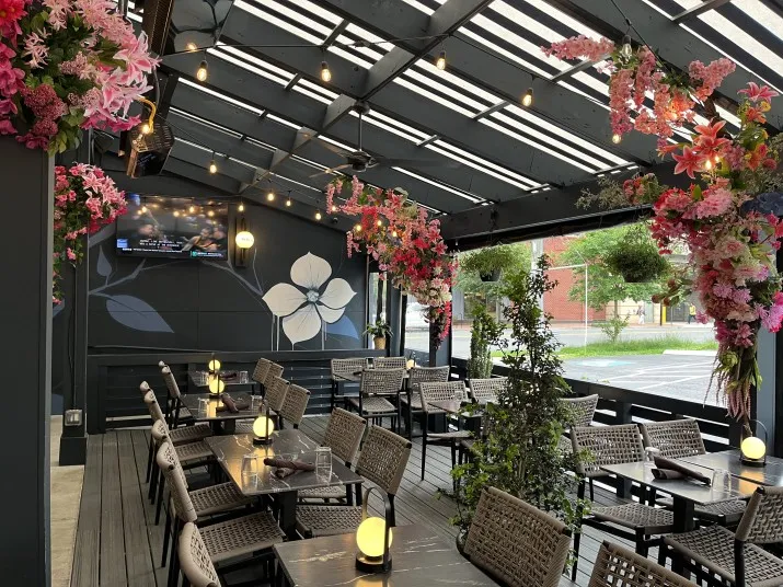 A covered outdoor dining area with tables and chairs, decorated with hanging pink flowers, greenery, and string lights. There is a mural of white flowers on the back wall and a TV mounted above.