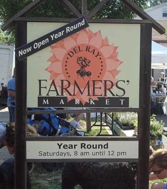 A sign for Del Ray Farmers Market reads Now Open Year Round and Saturdays, 8 am until 12 pm. People and tents are visible in the background, indicating an outdoor market setting.