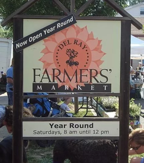 A sign for Del Ray Farmers Market reads Now Open Year Round and Saturdays, 8 am until 12 pm. People and tents are visible in the background, indicating an outdoor market setting.