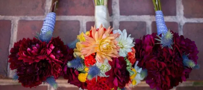 Three vibrant flower bouquets rest on red brick tiles. The central bouquet features a large peach dahlia surrounded by red, white, yellow, and blue blooms. The bouquets are wrapped with decorative ribbon at the stems.