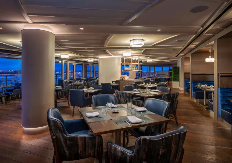 Modern, upscale restaurant interior with dark blue upholstered chairs, wooden tables set for dining, large windows offering city views at dusk, and soft ambient lighting.
